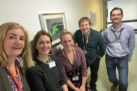 5 members of the Technician steering committee in a corridor at the Institute of Genetics and Cancer. On the wall behind them there is a picture of a genetically modified zebrafish embryo showing green fluorescent protein expression